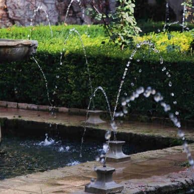 A classical stone fountain and arched water jets flow into a rectangular pond surrounded by manicured hedges and garden plants.