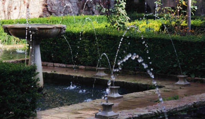 A classical stone fountain and arched water jets flow into a rectangular pond surrounded by manicured hedges and garden plants.
