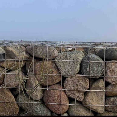A close-up view of a retaining wall built with wire mesh gabion baskets filled with large brown and grey fieldstones against an overcast sky.