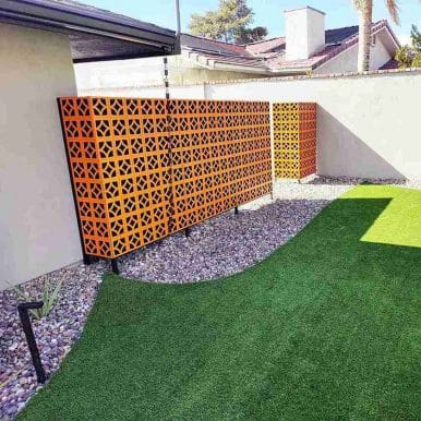 Orange geometric lattice privacy screen along a stucco wall in a sunny backyard with gravel and artificial turf.