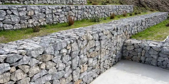 Tiered wire mesh gabion baskets filled with grey rocks installed along a sloped lawn for hill slope stabilization.